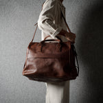 A person in a light-colored outfit carries a large brown leather duffel bag against a textured gray wall.