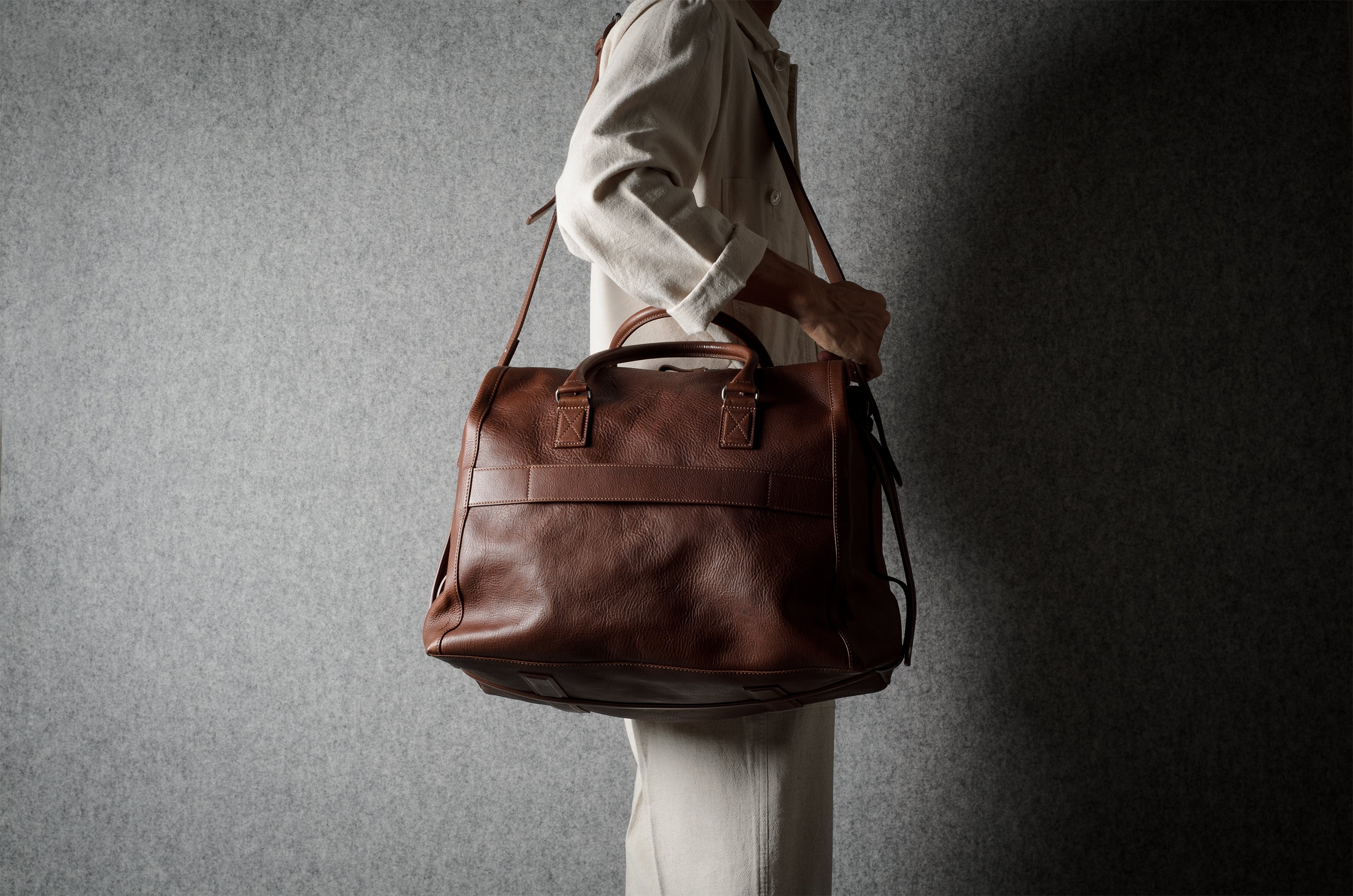 A person in a light-colored outfit carries a large brown leather duffel bag against a textured gray wall.