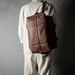 A person viewed from behind wearing a beige-colored outfit and carrying a brown leather bag against a textured gray background.