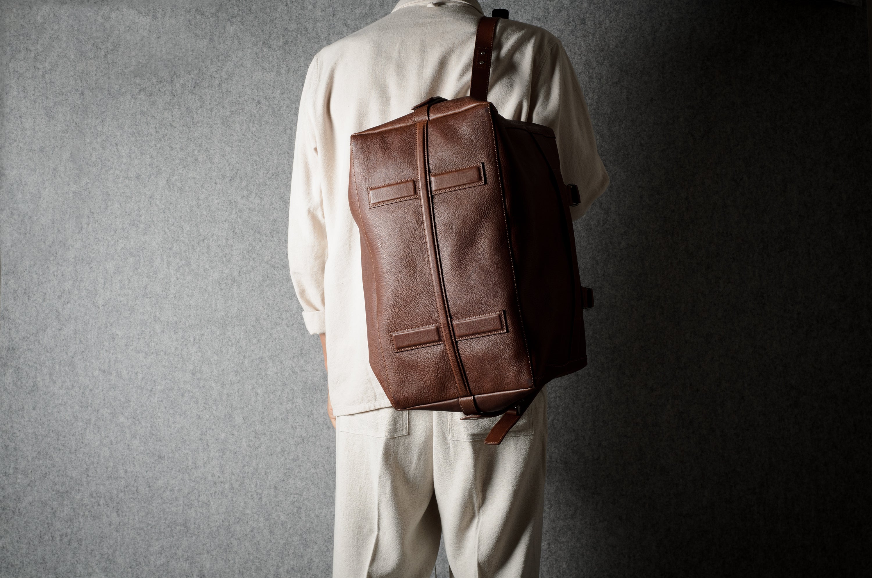 A person viewed from behind wearing a beige-colored outfit and carrying a brown leather bag against a textured gray background.
