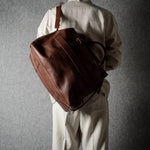 A person viewed from behind wearing a light beige outfit and carrying a large brown leather duffel bag over the shoulder against a textured gray background.