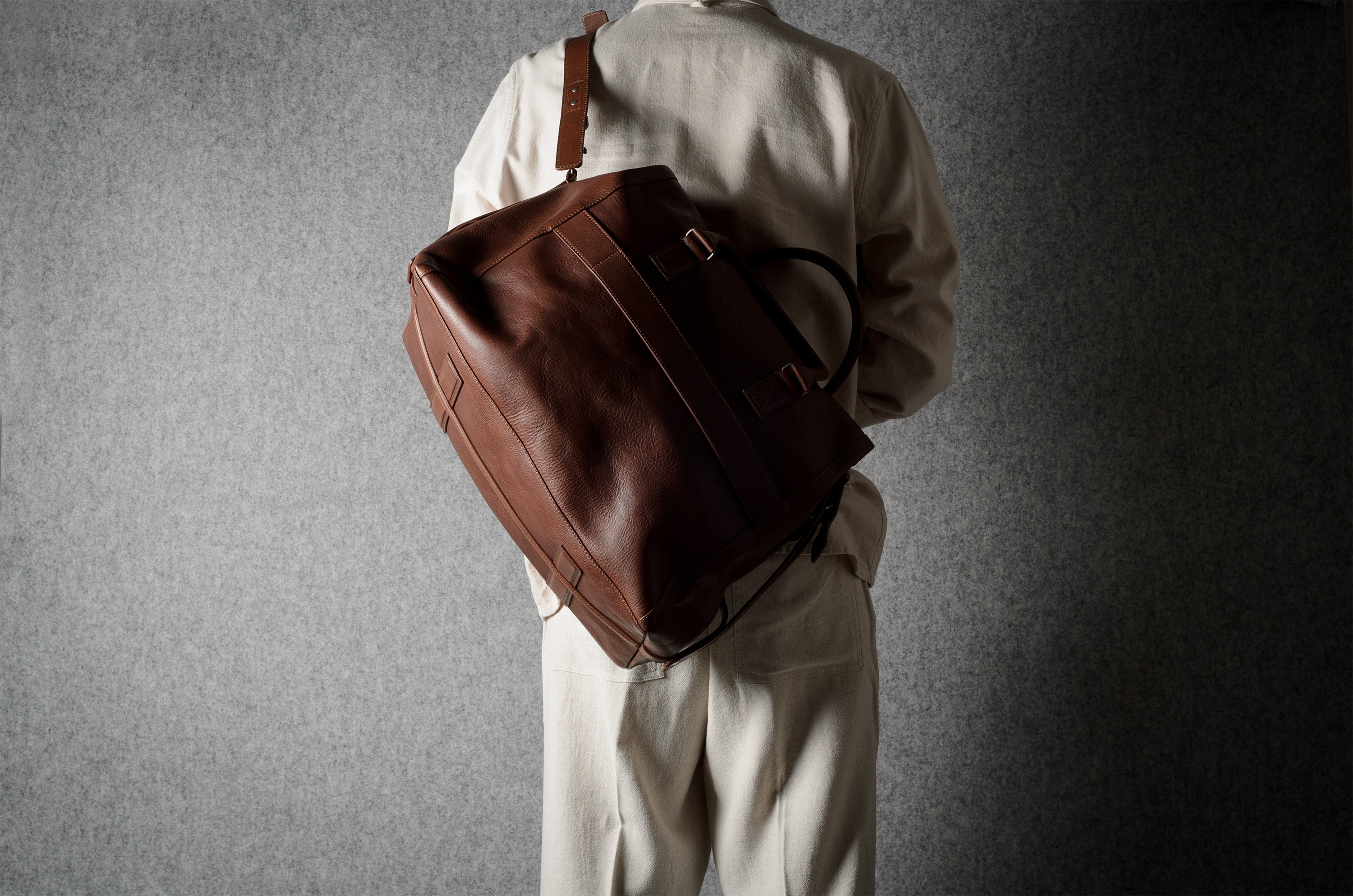 A person viewed from behind wearing a light beige outfit and carrying a large brown leather duffel bag over the shoulder against a textured gray background.