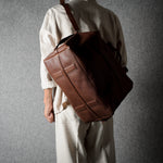 A person wearing a cream-colored traditional outfit carries a large brown leather duffel bag over their shoulder against a textured gray background.