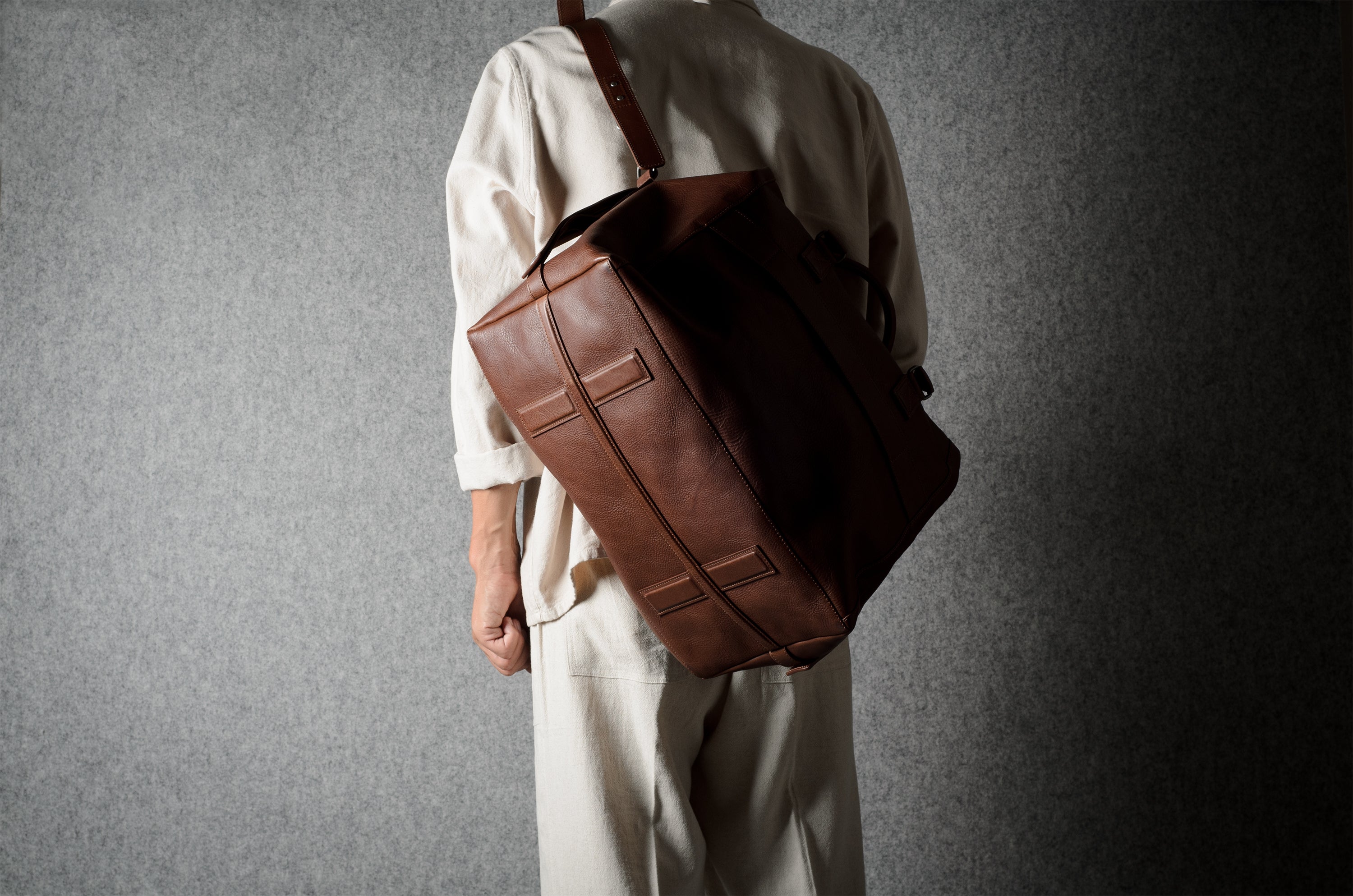 A person wearing a cream-colored traditional outfit carries a large brown leather duffel bag over their shoulder against a textured gray background.
