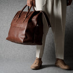 A person wearing beige trousers and tan shoes holds a large brown leather duffel bag against a textured gray backdrop.