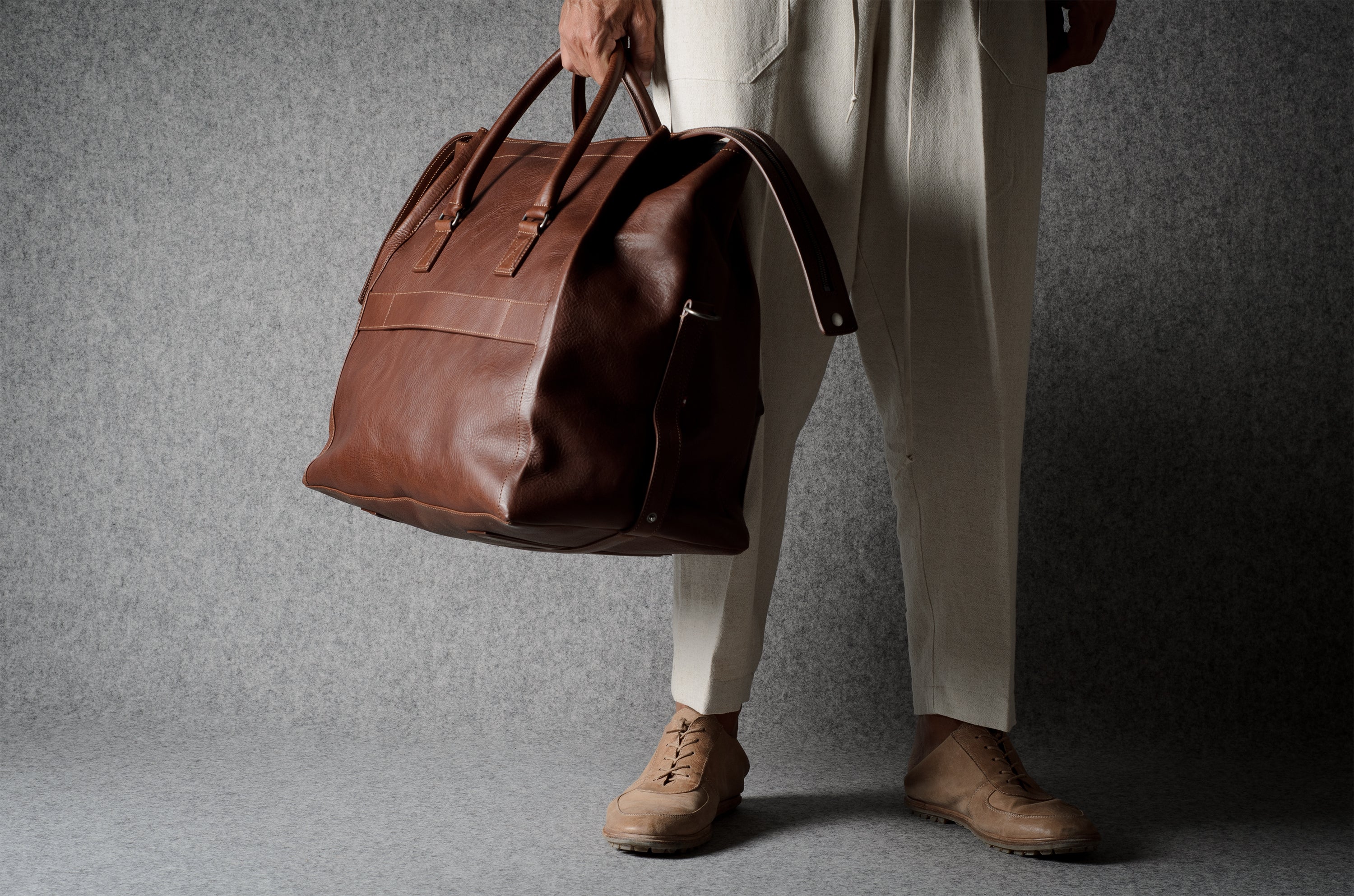 A person wearing beige trousers and tan shoes holds a large brown leather duffel bag against a textured gray backdrop.
