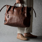 A person in beige trousers and tan shoes holds a large brown leather duffel bag against a textured gray background.