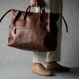 A person in beige trousers and tan shoes holds a large brown leather duffel bag against a textured gray background.