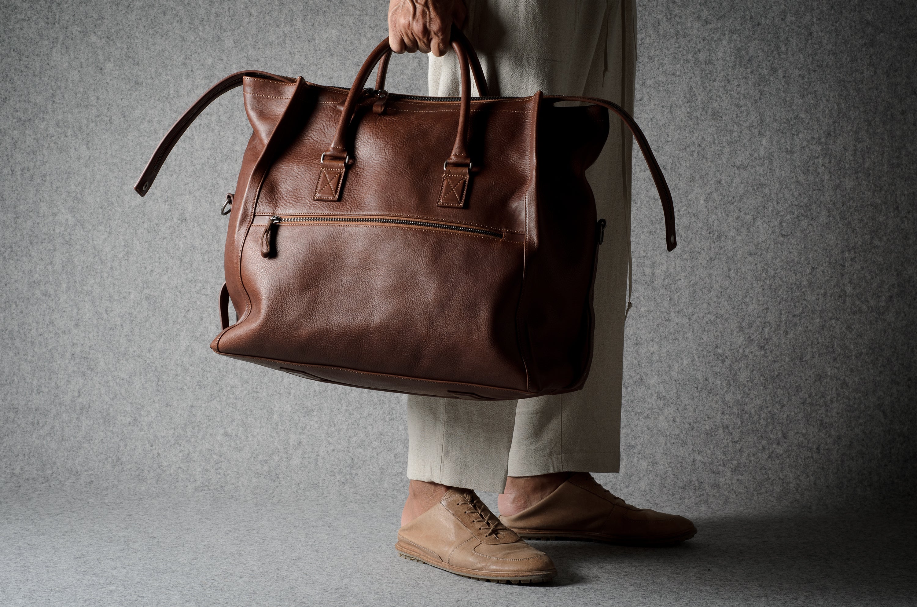 A person in beige trousers and tan shoes holds a large brown leather duffel bag against a textured gray background.