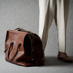 A brown leather duffel bag placed on a gray floor next to a person wearing cream trousers and brown shoes, photographed in a studio setting.