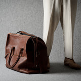 A brown leather duffel bag placed on a gray floor next to a person wearing cream trousers and brown shoes, photographed in a studio setting.