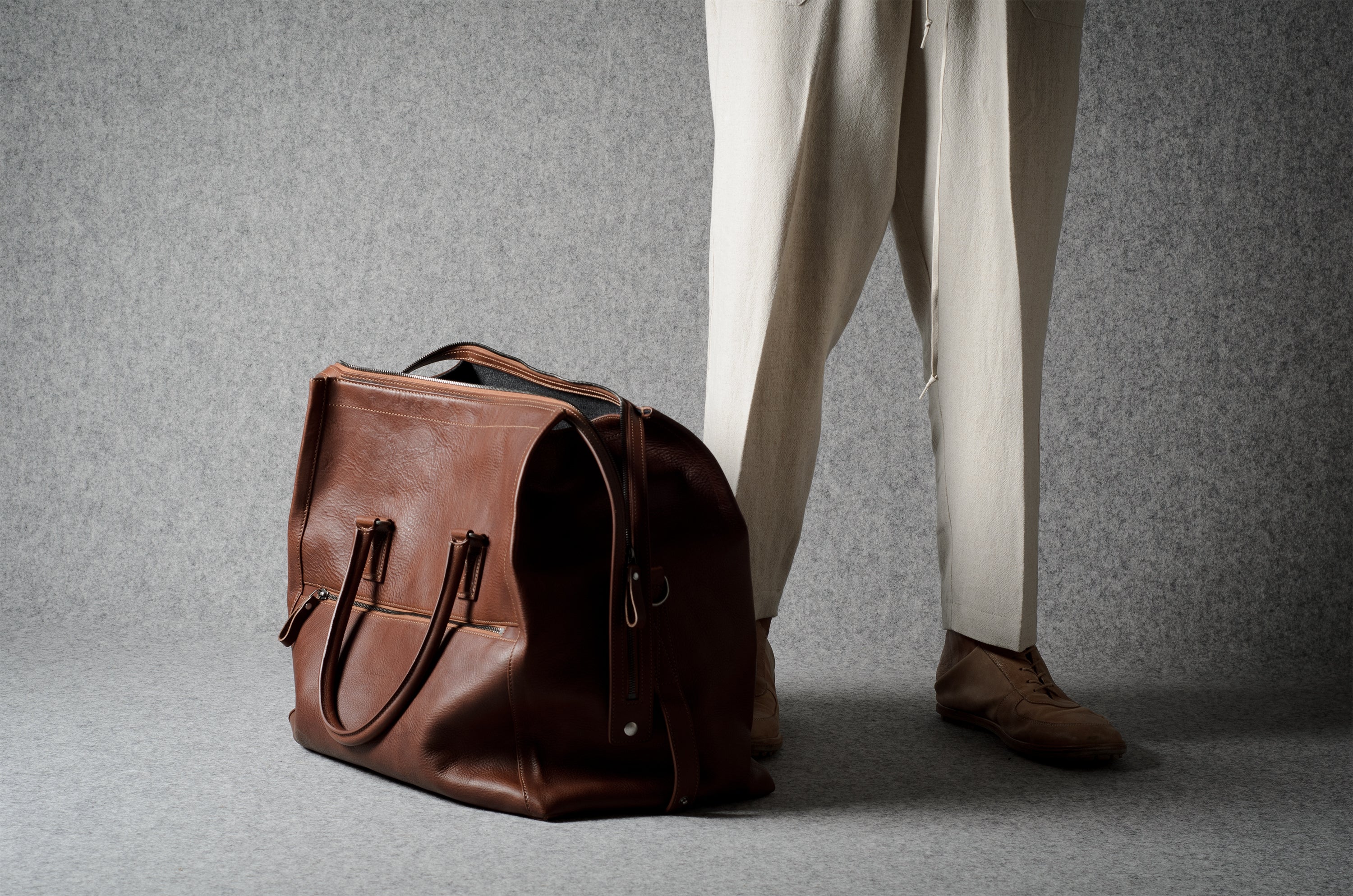 A brown leather duffel bag placed on a gray floor next to a person wearing cream trousers and brown shoes, photographed in a studio setting.