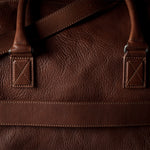 Close-up of a brown leather duffel bag showing the textured grain, double strap attachments, and stitching.