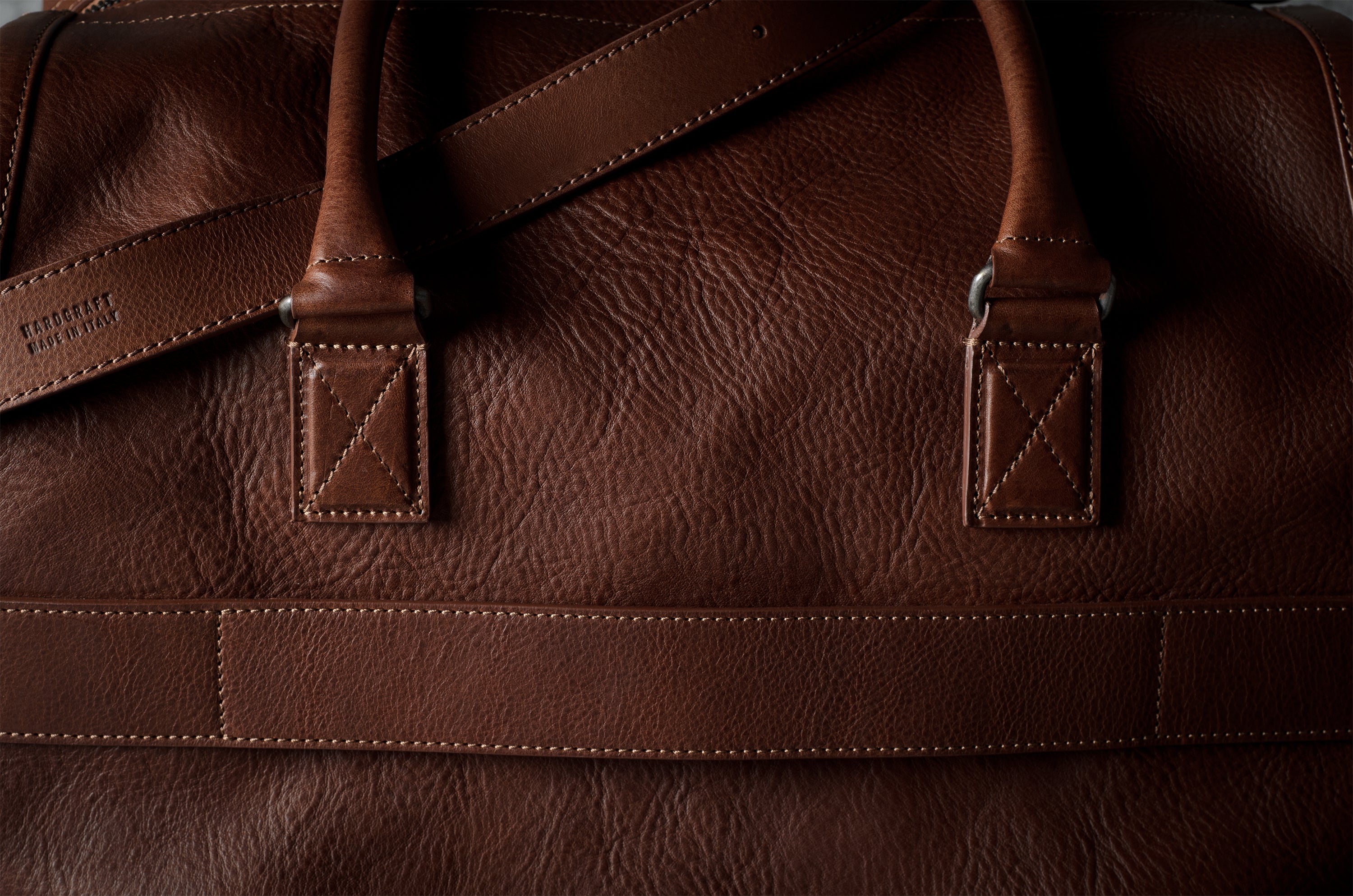 Close-up of a brown leather duffel bag showing the textured grain, double strap attachments, and stitching.