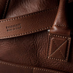 Close-up of a brown leather bag showing the textured grain, stitching, and an embossed brand mark on a leather strap.