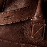 Close-up of a brown leather bag showing the textured grain, stitching, and an embossed brand mark on a leather strap.