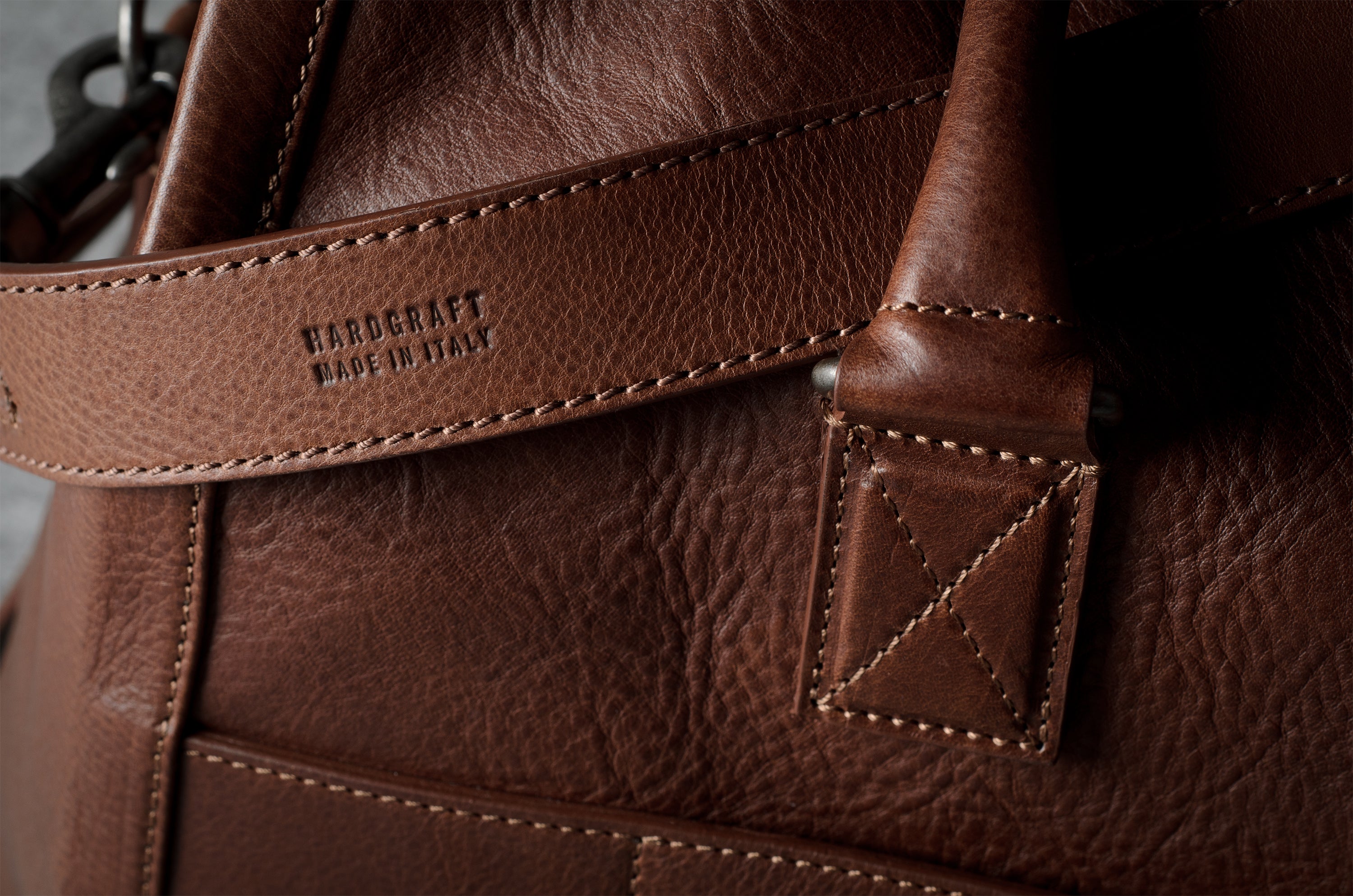 Close-up of a brown leather bag showing the textured grain, stitching, and an embossed brand mark on a leather strap.