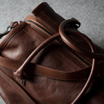 A close-up of a brown leather duffel bag with a natural grainy texture, featuring a zippered compartment and a thick leather handle, set against a textured gray background.
