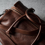 A close-up of a brown leather duffel bag with a natural grainy texture, featuring a zippered compartment and a thick leather handle, set against a textured gray background.