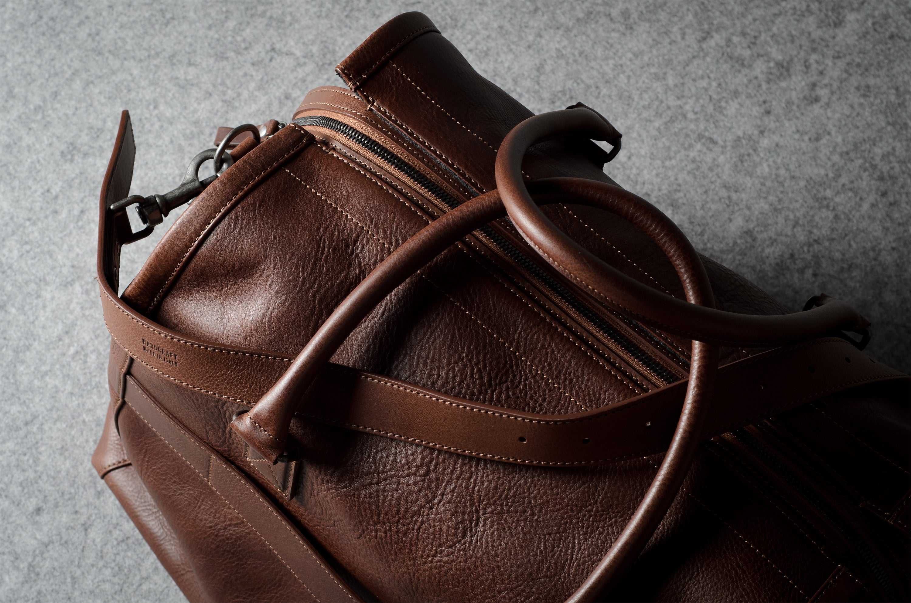 A close-up of a brown leather duffel bag with a natural grainy texture, featuring a zippered compartment and a thick leather handle, set against a textured gray background.