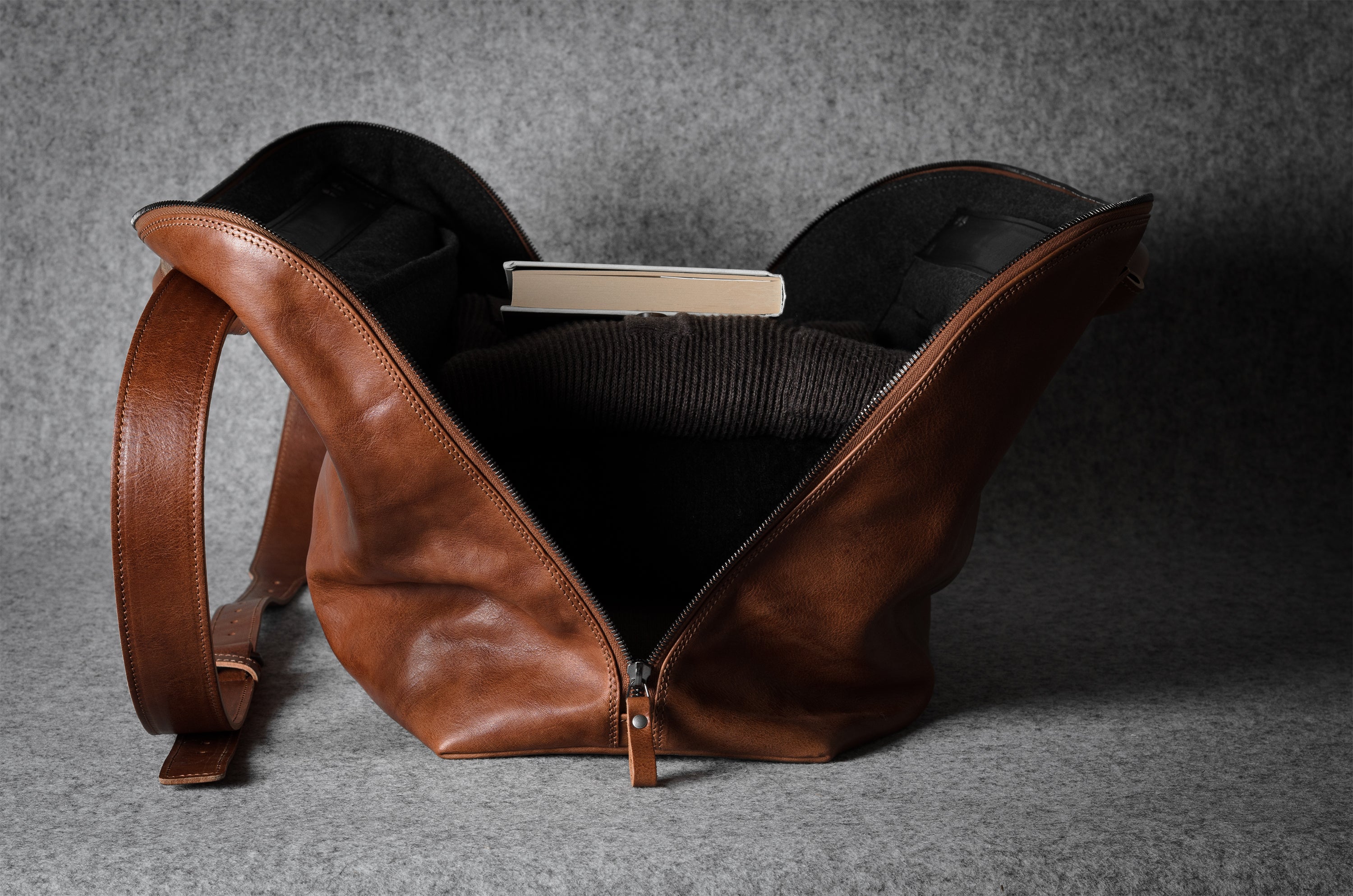 Brown leather bag with a grey interior, showing a sweater and book on a gray background
