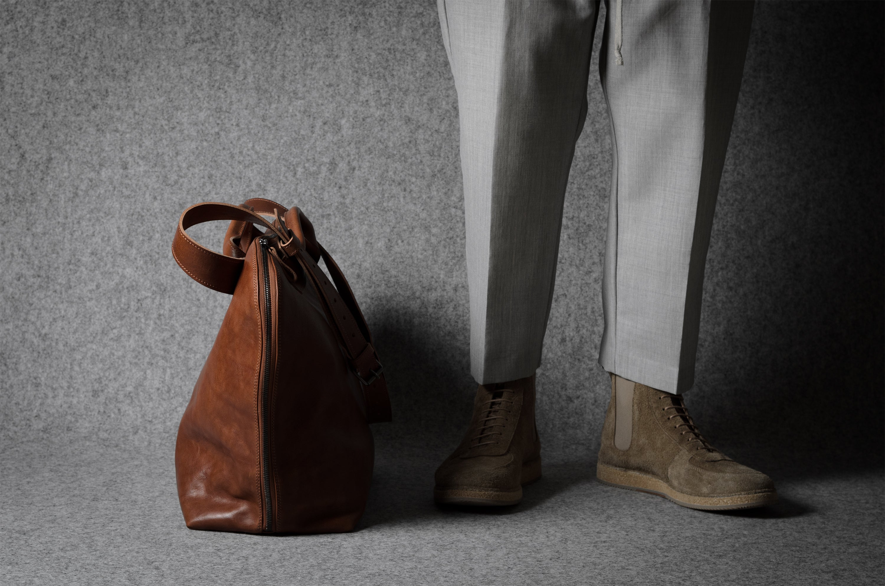 Brown leather bag next to a person wearing light gray pants and brown shoes on a gray background