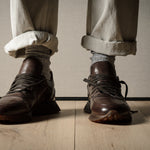Brown leather sneakers worn with rolled-up beige pants on a wooden floor.